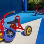 Bright red tricycles with yellow wheels parked on a sunlit concrete surface at Buttercups Perth Childcare. A blue helmet rests on one of the tricycles, highlighting the emphasis on safety. The area is partially shaded by a blue canopy, with greenery and a blue pole visible in the background, creating a vibrant and engaging environment for children.