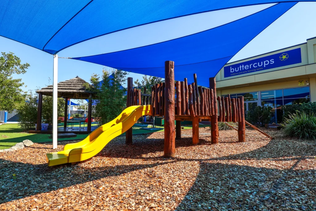 Outdoor play area at Buttercups Perth Childcare featuring a vibrant yellow slide and a wooden climbing structure, set amidst a natural mulch ground. The space is shaded with a large blue canopy, providing a sun-safe environment for children. A thatched gazebo and greenery in the background add to the inviting, nature-inspired setting.