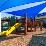 Outdoor play area at Buttercups Perth Childcare featuring a vibrant yellow slide and a wooden climbing structure, set amidst a natural mulch ground. The space is shaded with a large blue canopy, providing a sun-safe environment for children. A thatched gazebo and greenery in the background add to the inviting, nature-inspired setting.