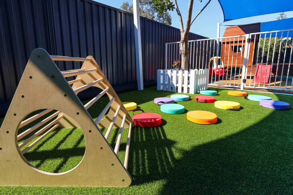 An engaging play area at Buttercups Perth Childcare featuring a wooden climbing frame and colourful stepping stones arranged on vibrant green grass. The space is shaded and fenced, creating a safe and stimulating environment for children to explore and develop their motor skills.