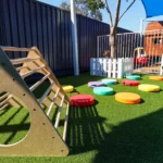 An engaging play area at Buttercups Perth Childcare featuring a wooden climbing frame and colourful stepping stones arranged on vibrant green grass. The space is shaded and fenced, creating a safe and stimulating environment for children to explore and develop their motor skills.