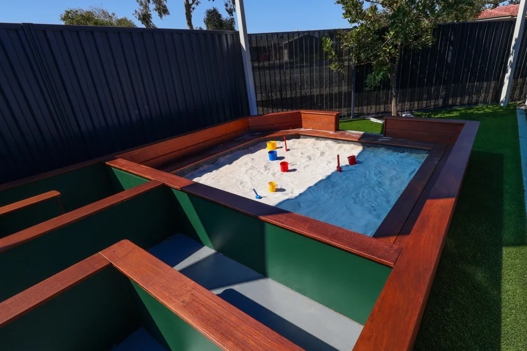 A well-designed sandpit area at Buttercups Perth Childcare, featuring wooden framing and equipped with colourful buckets and spades. The sandpit is surrounded by green spaces, offering children a safe and inviting environment to explore and play outdoors.