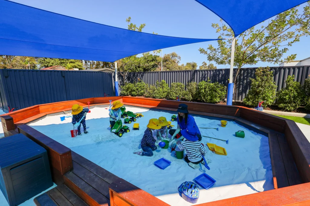 Children and a teacher enjoying activities in a shaded sandpit area at Buttercups Perth Childcare. The children, wearing yellow hats, play with toys like trucks, buckets, and spades under vibrant blue sunshades, fostering creativity and engagement in a safe outdoor environment surrounded by greenery.