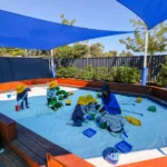 Children and a teacher enjoying activities in a shaded sandpit area at Buttercups Perth Childcare. The children, wearing yellow hats, play with toys like trucks, buckets, and spades under vibrant blue sunshades, fostering creativity and engagement in a safe outdoor environment surrounded by greenery.