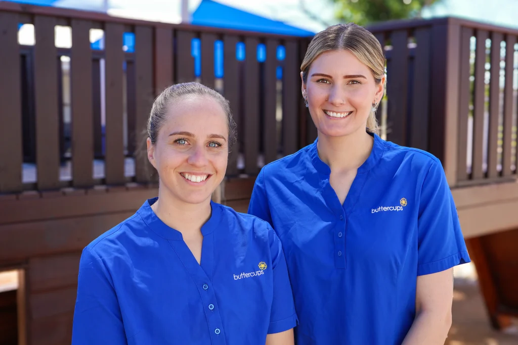 Buttercups Perth Childcare Hammond Park staff members, wearing blue uniforms, standing in front of wooden fort in the outdoor play area, showcasing their welcoming and professional environment.