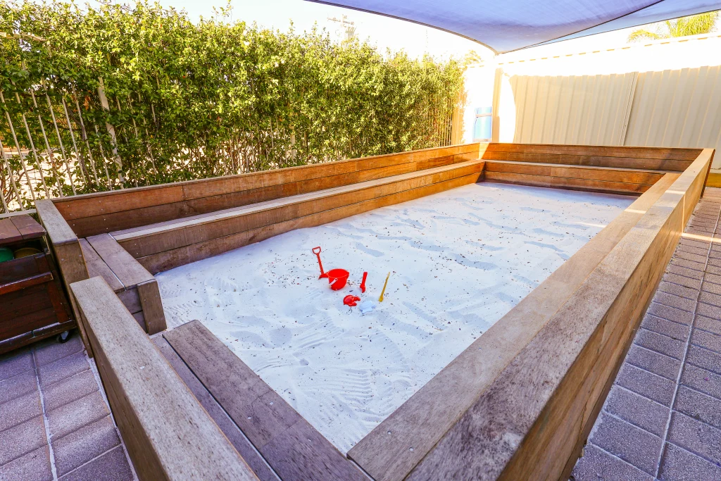 Large outdoor sandpit at Buttercups Childcare, enclosed by sturdy wooden seating for group play and social interaction. The bright white sand features scattered red and yellow sand toys, inviting children to engage in imaginative and sensory play.