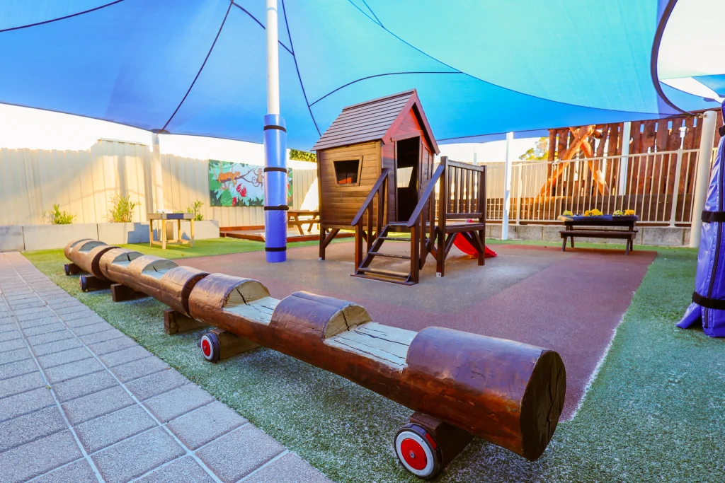 Shaded outdoor play area at Buttercups Perth Childcare featuring a wooden cubby house with a slide, a natural log balancing beam on wheels, soft fall rubber flooring, and colourful murals. The secure, well-maintained space encourages imaginative and active play in a safe environment.