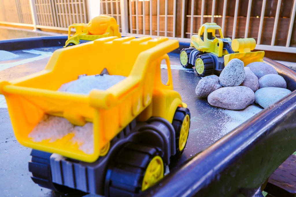 Close-up of a sensory play table at Buttercups Perth Childcare, featuring yellow toy construction vehicles, including a dump truck, bulldozer, and cement mixer. The setup includes real rocks and sand, encouraging hands-on, imaginative play and fine motor skill development in a secure outdoor environment.