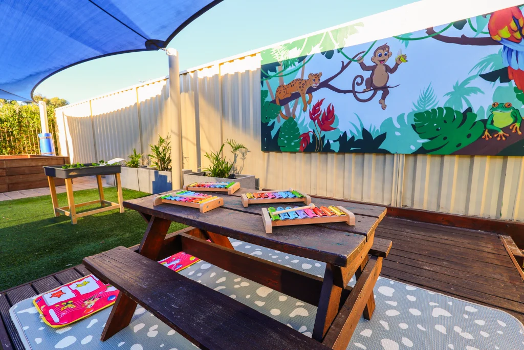 Shaded outdoor learning space at Buttercups Perth Childcare featuring a wooden picnic table with colourful xylophones and a musical play mat. A jungle-themed mural and raised garden beds enhance the vibrant, nature-inspired setting, encouraging creativity and sensory exploration in a secure environment.