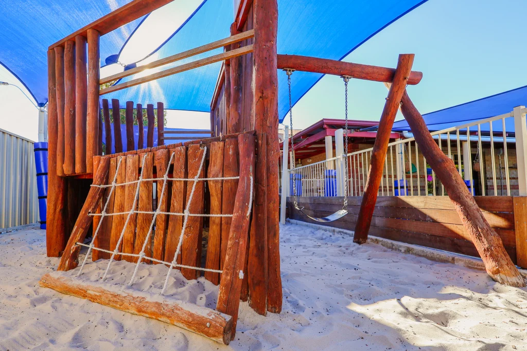 Natural wooden outdoor play structure at Buttercups Childcare, featuring a climbing net, a platform with railings, a swing, and soft sand beneath for safe play. The area is shaded with blue sails, creating a welcoming and sun-safe environment for children to explore and develop their physical skills.