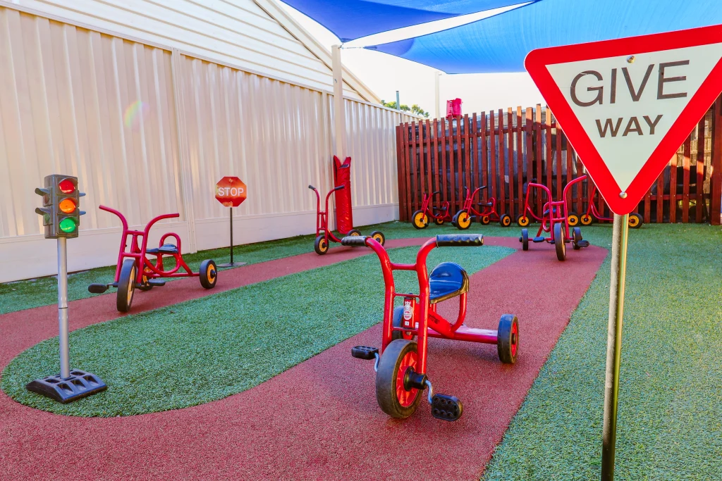 Outdoor bike track at Buttercups Childcare featuring red tricycles, realistic road signs, and a traffic light to create an engaging and educational environment. The track is made of soft, rubberised material and surrounded by artificial grass, with shaded areas to ensure sun-safe play.