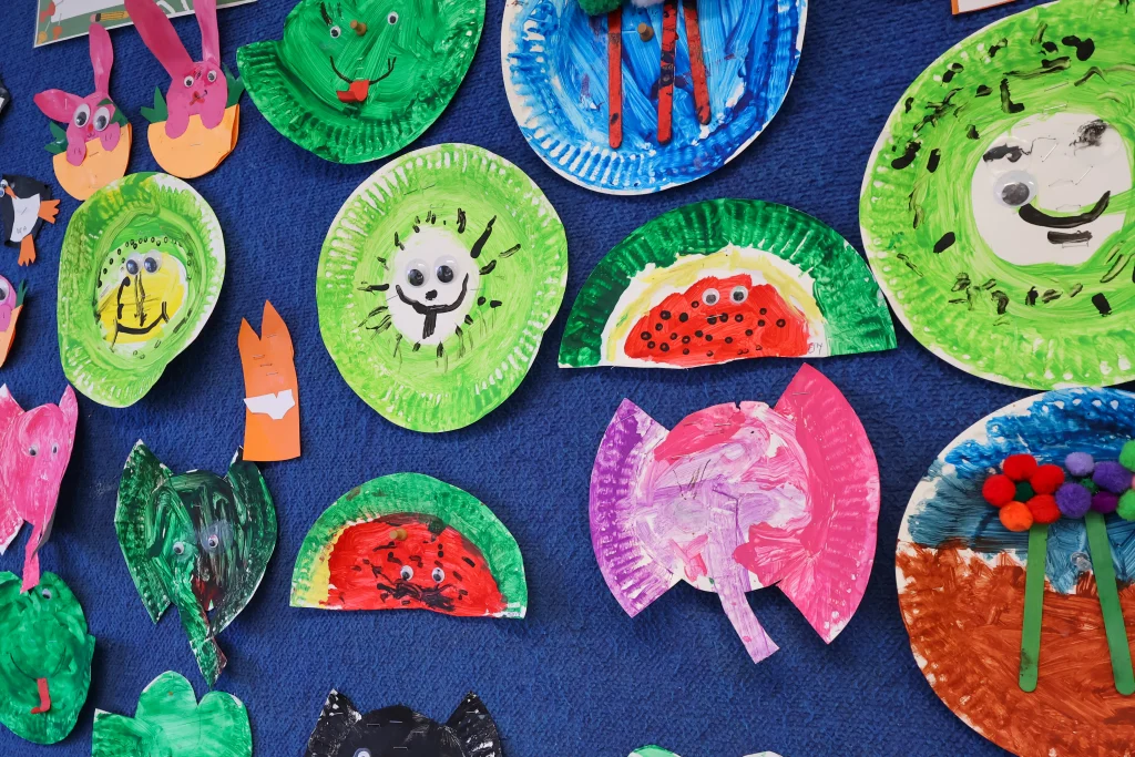 A vibrant "Art of the Month" display at Buttercups Childcare, showcasing children's creative paper plate artwork on a deep blue background. The mix of bright colors, textures, and materials highlights the children's creativity and fine motor skill development.