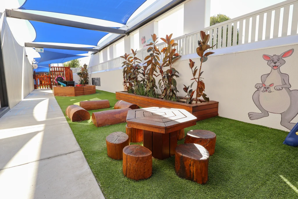 A natural outdoor baby play area at Buttercups Childcare with wooden play elements, artificial grass, and shaded seating. Raised garden beds and a kangaroo mural add a playful, Australian touch.