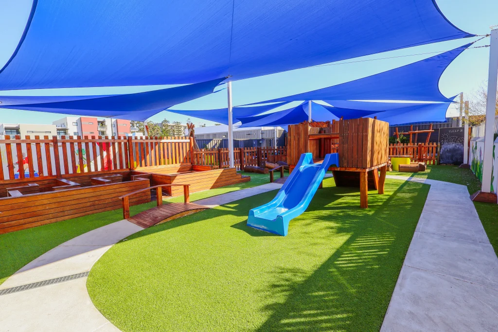 The outdoor play area at Buttercups Perth Childcare features a wooden play structure with a blue slide shaded by large blue sails.