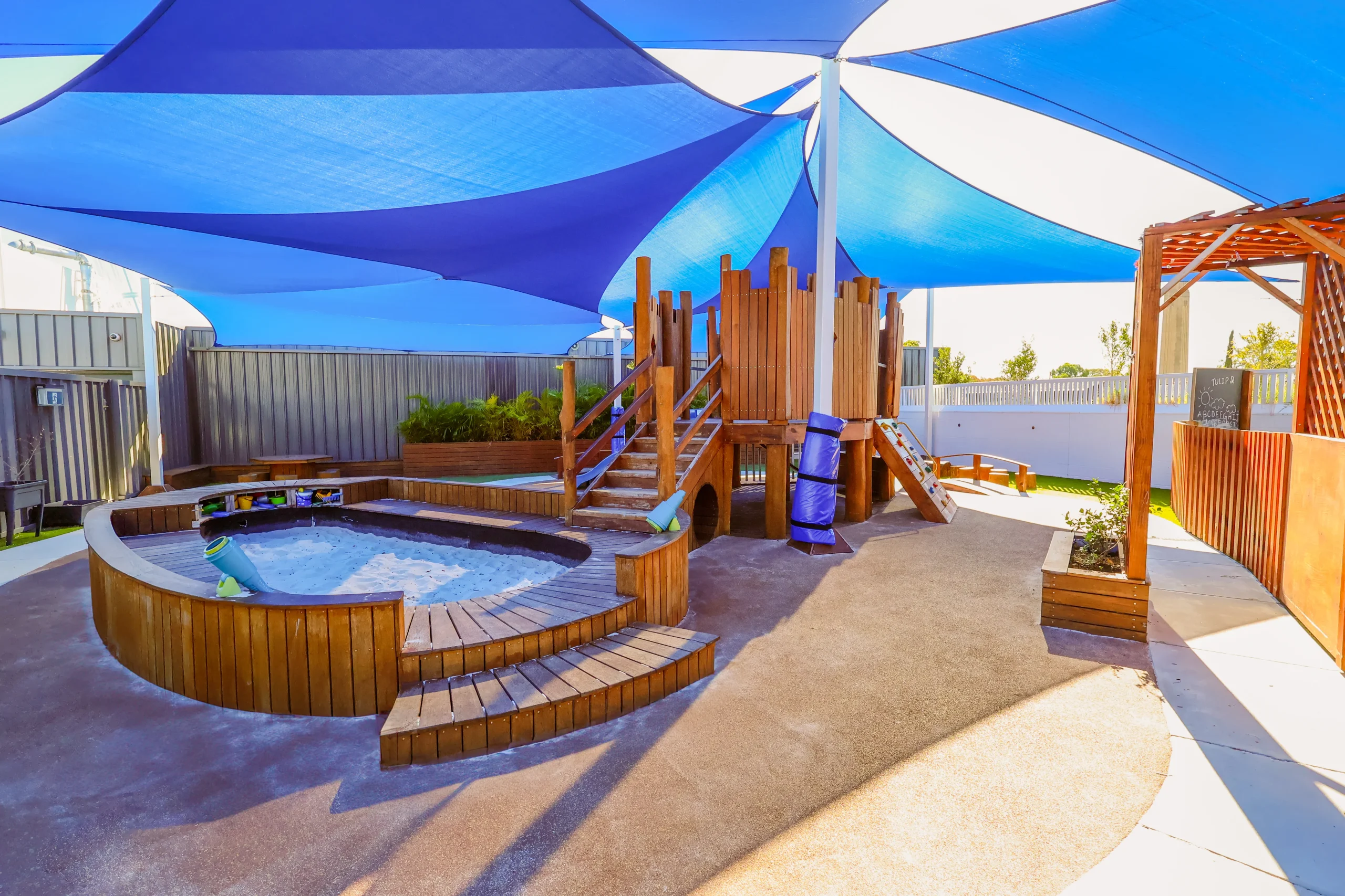 The modern outdoor play area at Buttercups Perth Childcare features a wooden play structure and seating area, and it is shaded by blue and white sails with sunlight streaming through.