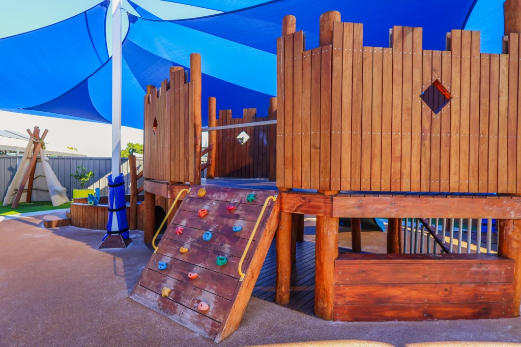 A wooden fort-style playground at Buttercups Childcare featuring a climbing wall, shaded play areas, and a teepee in the background. The natural design encourages imaginative and active play.