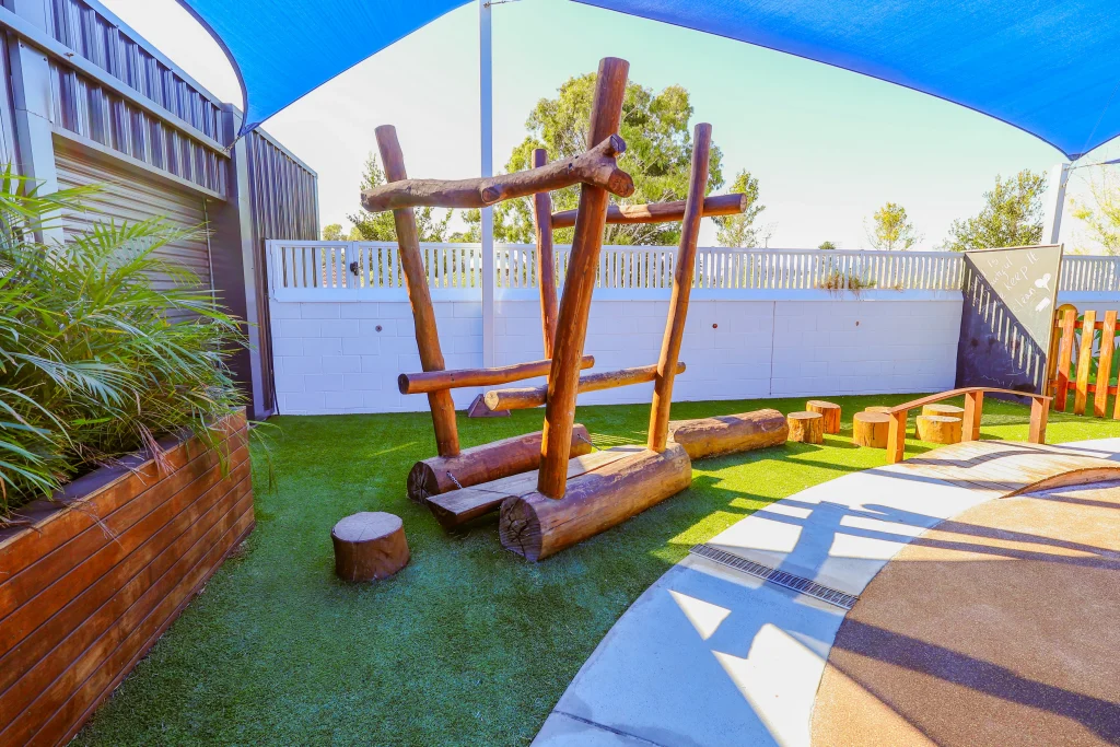 The modern outdoor play area at Buttercups Perth Childcare features a natural wood climbing frame with a balancing beam and wooden stepping stones. This inviting play area encourages children to develop balance and coordination in a shaded, outdoor setting.