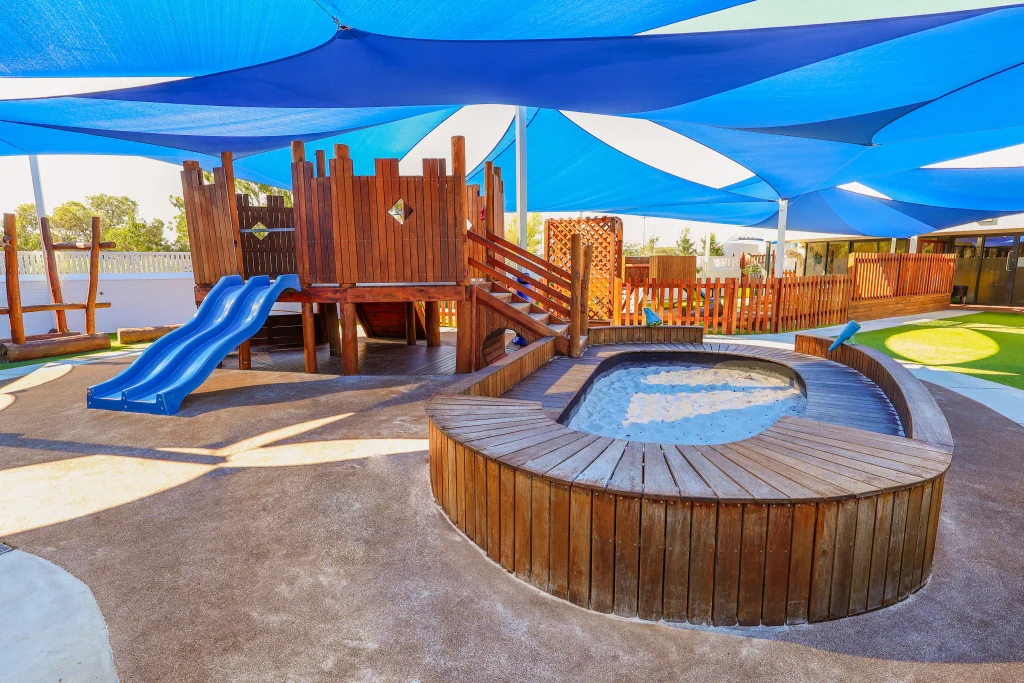 The wooden play structure with twin blue slides at Buttercups Perth Childcare is shaded by blue and white sails, offering a safe and fun environment for children.