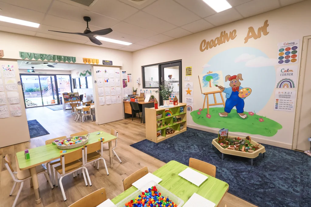 The bright and airy kindy room at Buttercups Perth Childcare features interactive activities on each table. A charming wall mural of a rabbit painting an outdoor scene adds a playful and creative touch to the space.