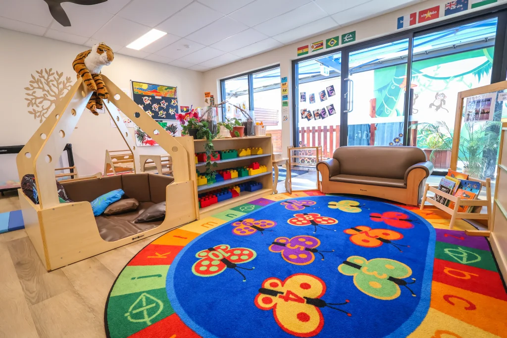 The vibrant toddler play space at Buttercups Perth Childcare is designed for learning and fun. It features a cosy reading nook, building blocks, and a butterfly-themed rug with numbers and letters, all illuminated by large windows overlooking the outdoor play area.