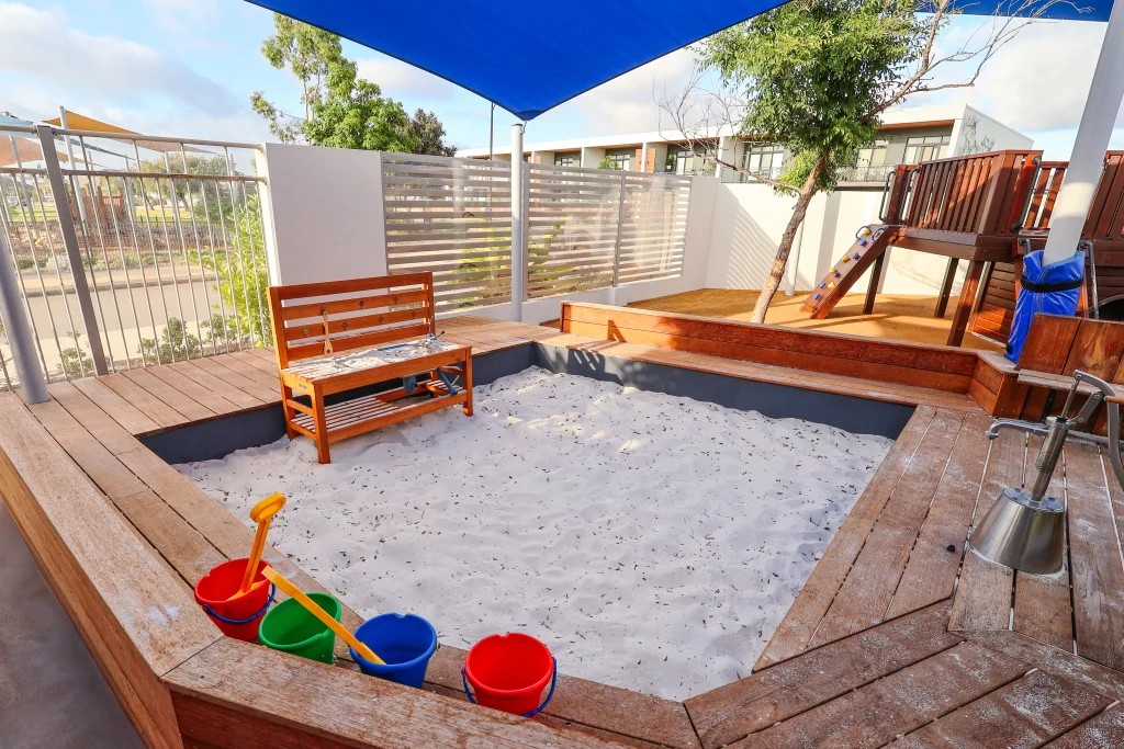 The modern outdoor play area at Buttercups Perth Childcare features a wooden play structure and seating area, and it is shaded by blue and white sails with sunlight streaming through.