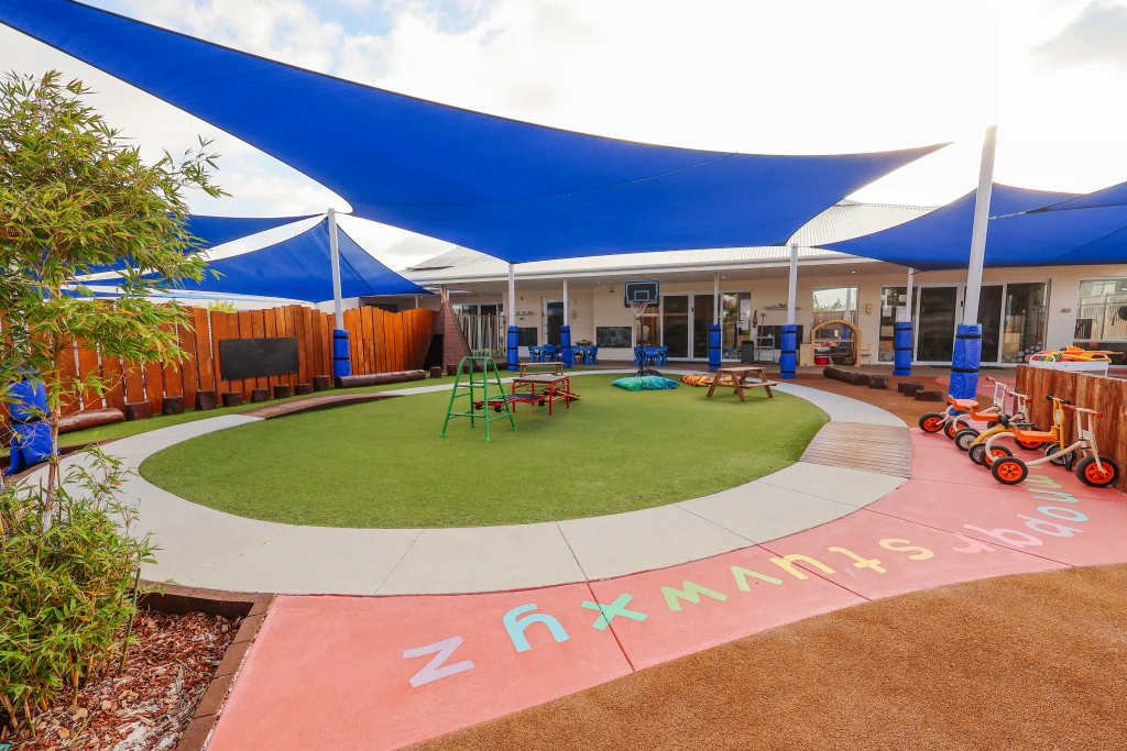 The spacious outdoor play area at Buttercups Perth Childcare features a large artificial grass oval surrounded by a dedicated bike track. With picnic benches, climbing toys, and balancing logs, this dynamic space encourages active play and social interaction. Blue shade sails provide a cool and comfortable environment for children to explore and enjoy.