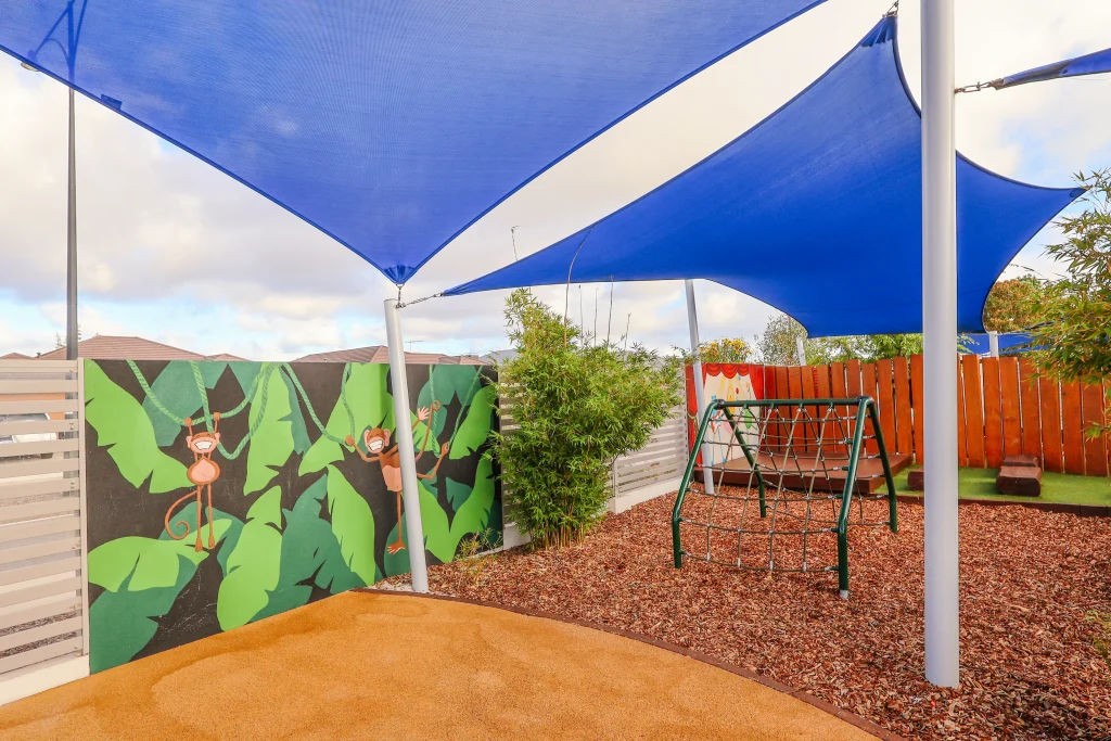 The vibrant outdoor play space at Buttercups Perth Childcare features a playful corner with a mural of banana leaves and monkeys, sparking children’s imagination. A sturdy climbing frame and a podium with logs in the background provide opportunities for active exploration. Blue shade sails offer a cool and inviting environment for outdoor adventures.