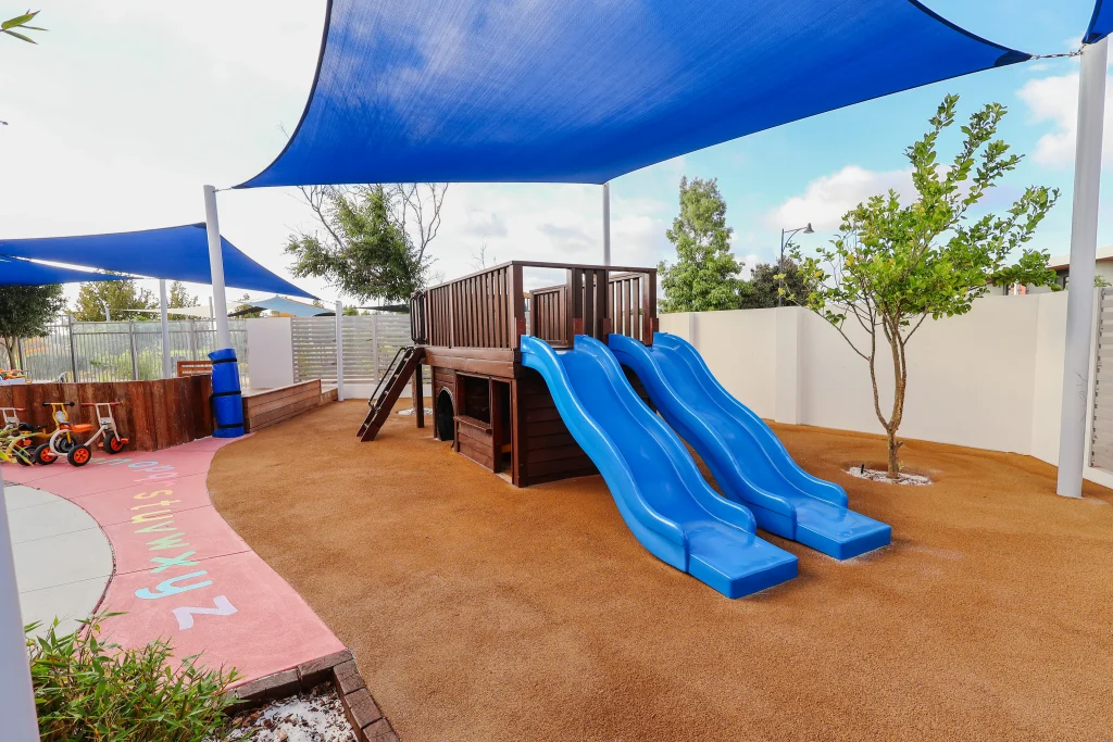 The wooden play structure with twin blue slides at Buttercups Perth Childcare is shaded by blue and white sails, offering a safe and fun environment for children.