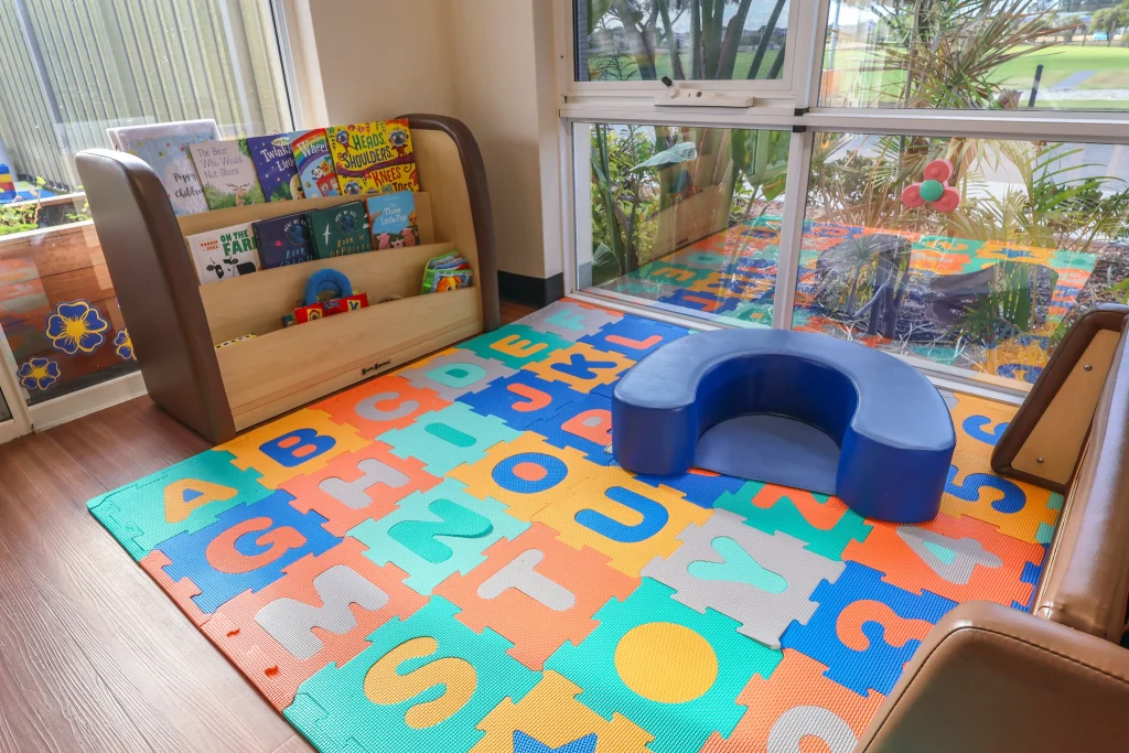 The cosy babies' reading corner at Buttercups Perth Childcare features colourful letter soft play mats and a supportive floor seat, providing a comfortable space for infants to engage in storytime with an educator. Age-appropriate bookshelves are stocked with vibrant children's books, encouraging early literacy and a love for reading in a warm and inviting environment.