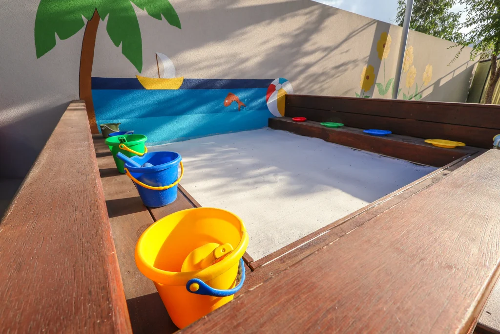 The inviting outdoor sandpit at Buttercups Perth Childcare is designed especially for babies, featuring a wooden structure with a built-in seating area around the edge. Colourful buckets and a vibrant mural add to the engaging play environment, encouraging sensory exploration and imaginative play.