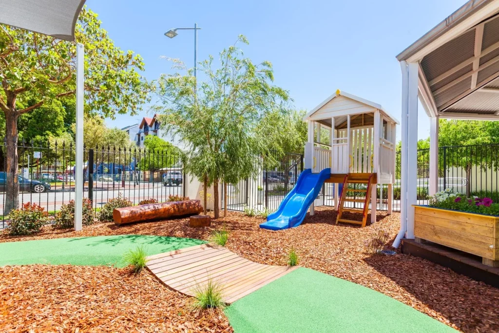 Outdoor play area at a Perth childcare centre, featuring a wooden cubby house with a blue slide, natural landscaping, and pathways for exploration.