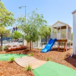 Outdoor play area at a Perth childcare centre, featuring a wooden cubby house with a blue slide, natural landscaping, and pathways for exploration.