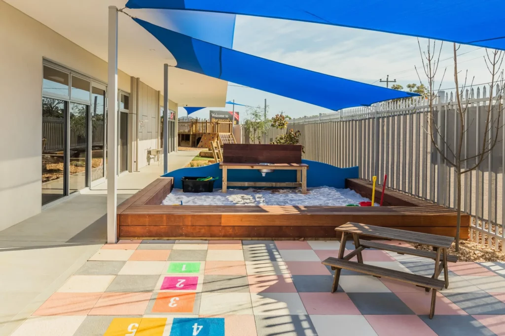 Outdoor play area at a Perth childcare centre, featuring a shaded sandpit, hopscotch tiles, and seating for creative and social play.