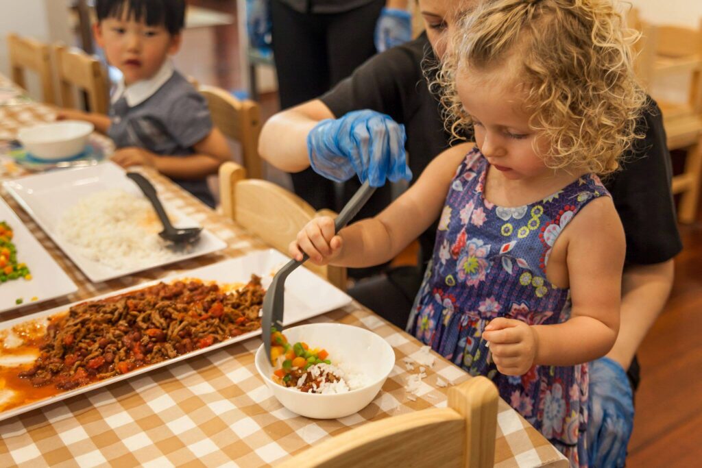 A child at Buttercups Perth Childcare learning independence by feeding herself lunch, with the gentle guidance of a staff member. The meal is nutritious, promoting healthy eating habits and self-confidence.