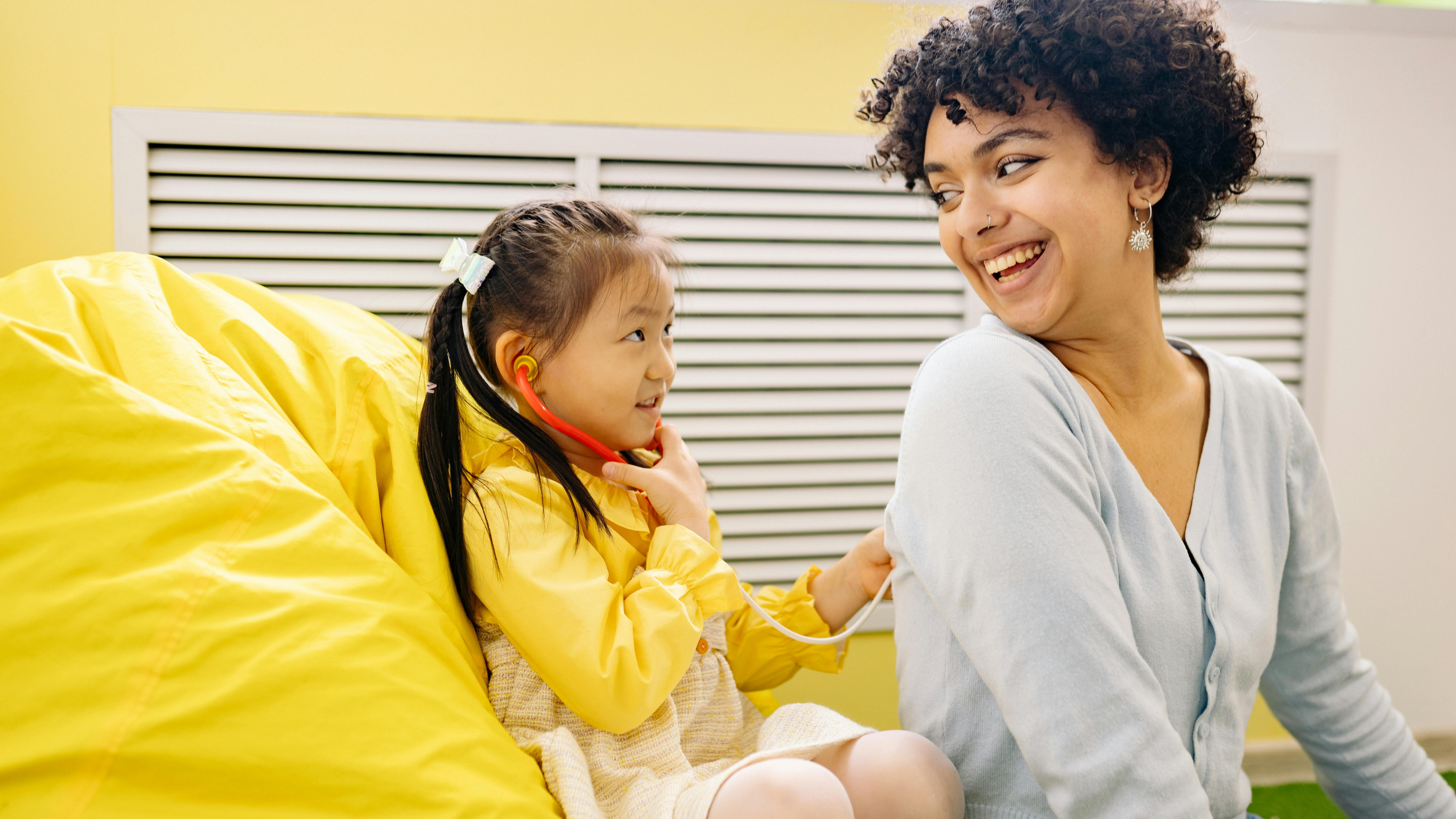 A woman with curly hair and a little girl engage in a doctor role-play, smiling as they engage in imaginative play at home.