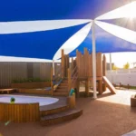 The modern outdoor play area at Buttercups Perth Childcare features a wooden play structure and seating area, and it is shaded by blue and white sails with sunlight streaming through.