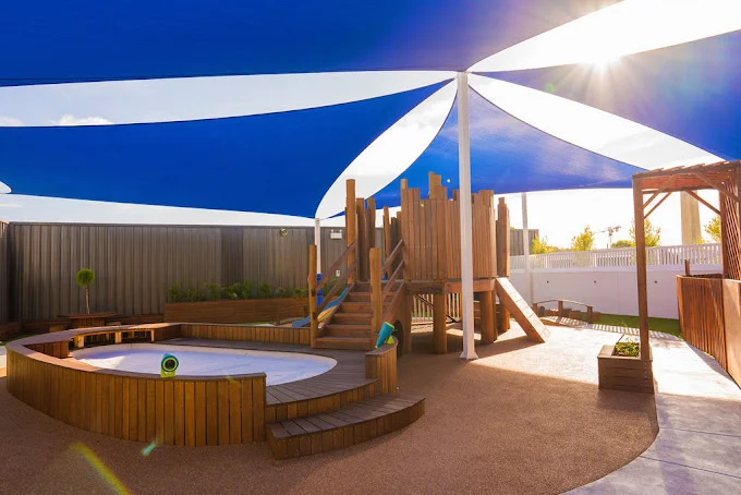 The modern outdoor play area at Buttercups Perth Childcare features a wooden play structure and seating area, and it is shaded by blue and white sails with sunlight streaming through.