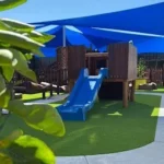 The outdoor play area at Buttercups Perth Childcare features a wooden play structure with a blue slide shaded by large blue sails.