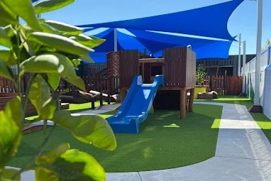 The outdoor play area at Buttercups Perth Childcare features a wooden play structure with a blue slide shaded by large blue sails.