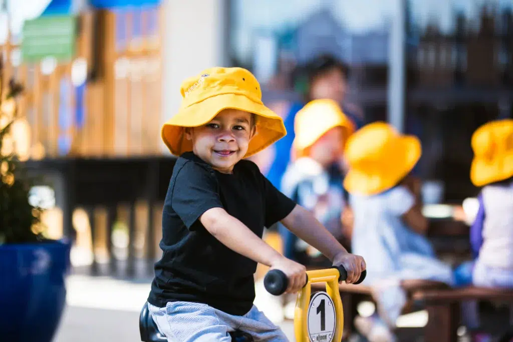 A cheerful child at Buttercups Perth Childcare rides a yellow tricycle, wearing a bright yellow sunhat and a playful smile. The outdoor play area buzzes with energy as other children engage in activities in the background, showcasing a vibrant and nurturing environment for early learning and fun.