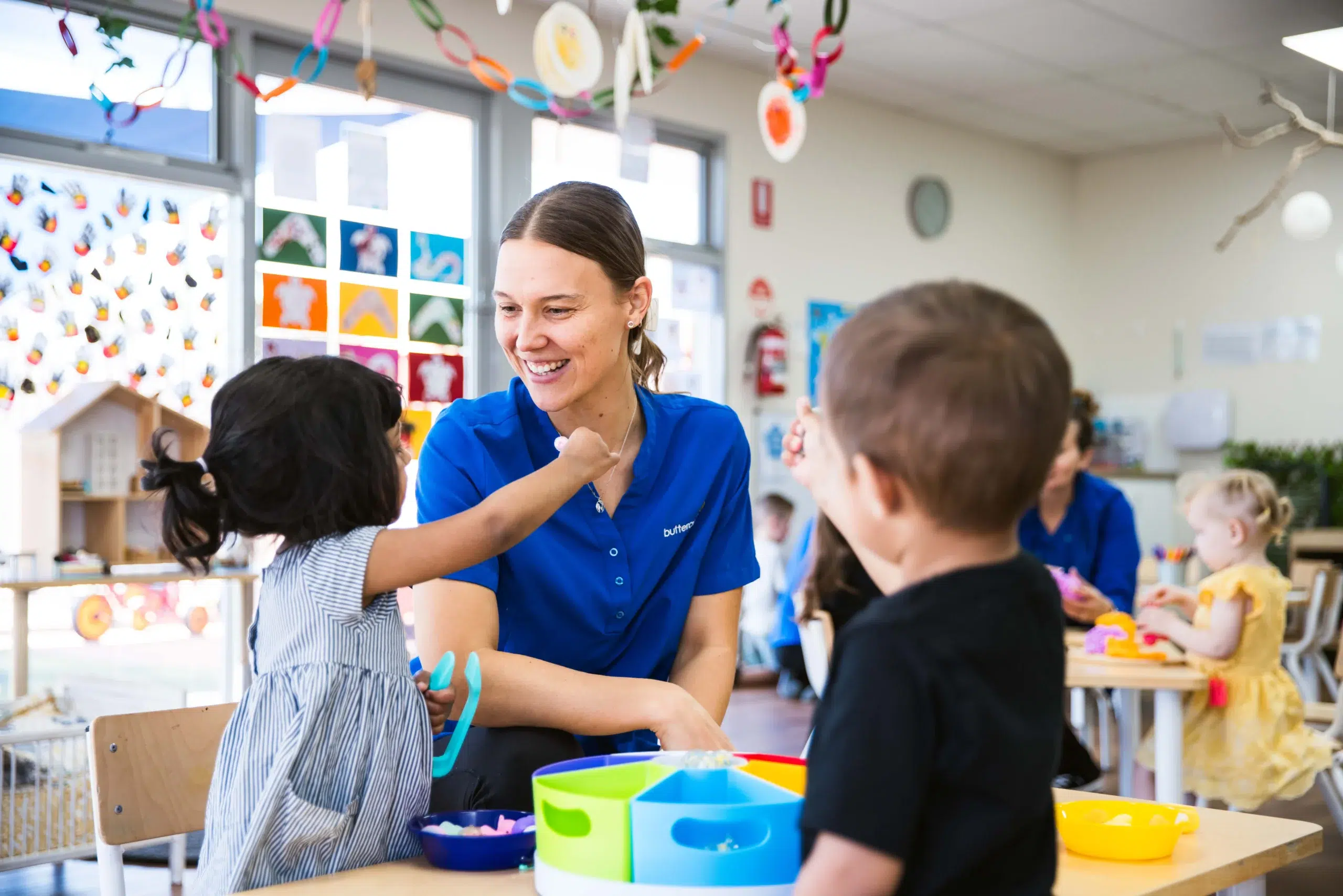 A joyful and engaging learning environment at Buttercups Perth Childcare, where a smiling educator in a blue uniform interacts with young children during a fun activity. The brightly decorated classroom features colourful artwork, playful decorations, and children engaging in hands-on learning, fostering creativity and connection.