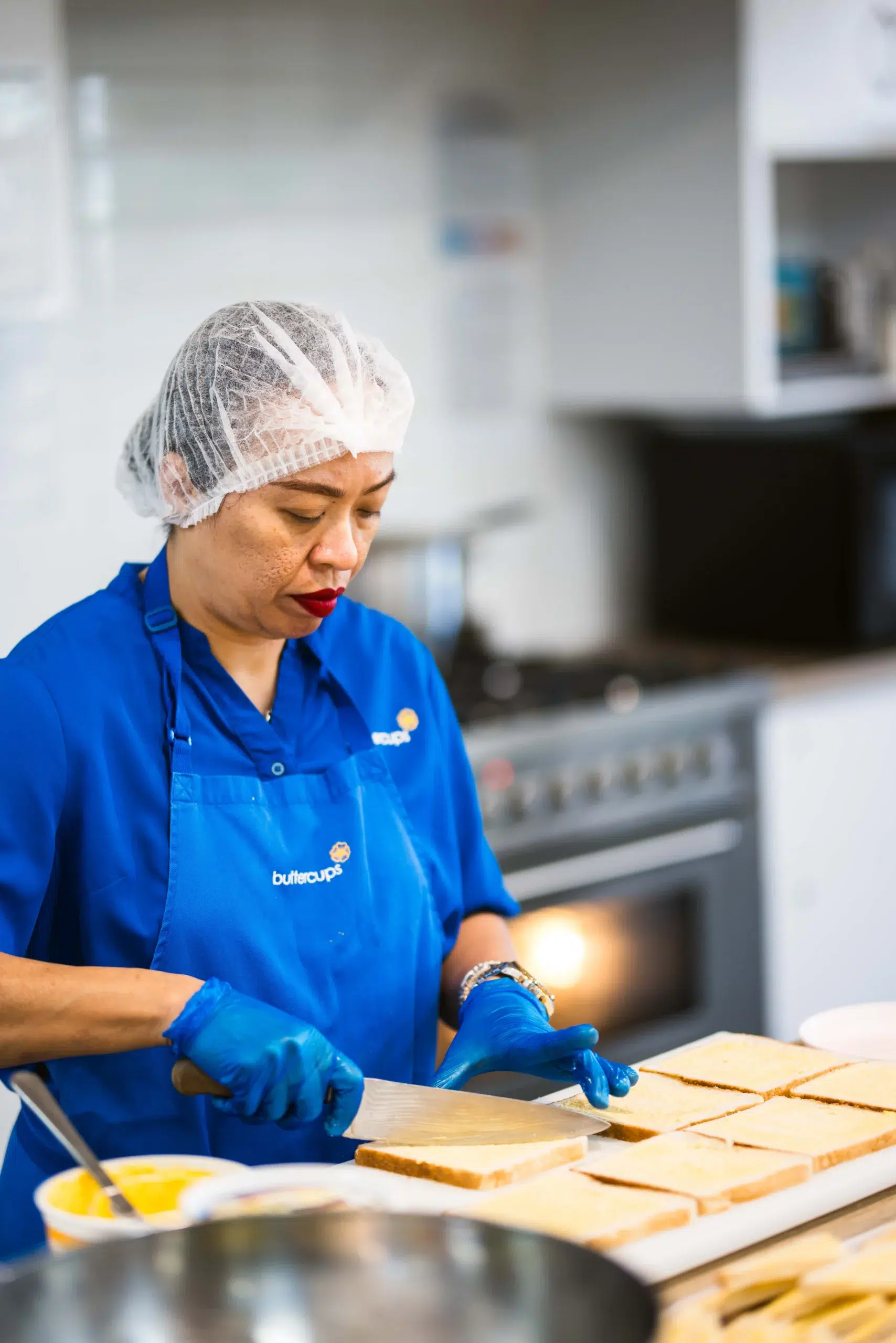 A dedicated staff member at Buttercups Perth Childcare, wearing a blue uniform, gloves, and a hairnet, carefully preparing sandwiches in a clean and well-equipped kitchen, ensuring nutritious meals for the children.