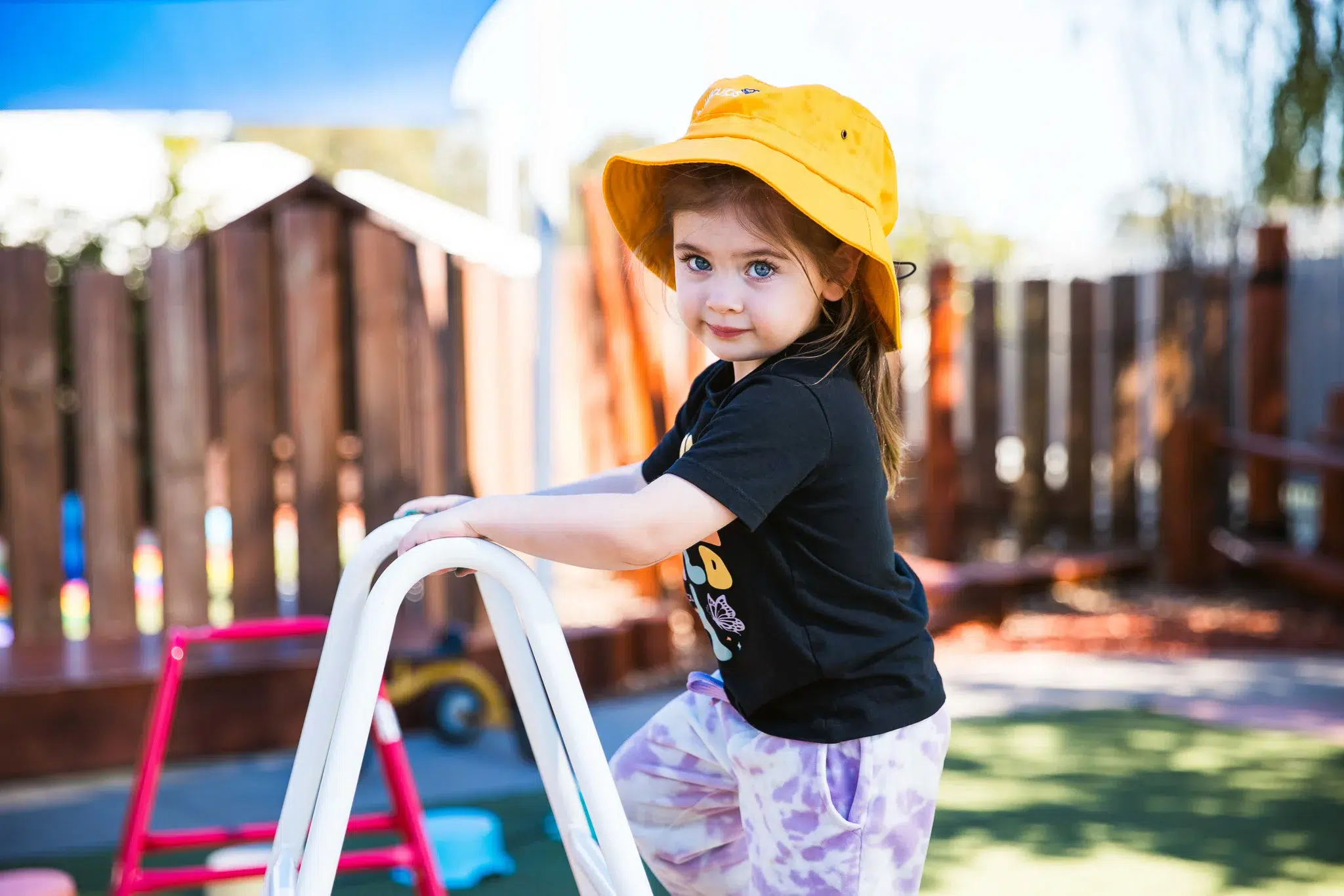 A cheerful child in a bright yellow hat eagerly climbs a ladder at Buttercups Perth Childcare, embracing adventure and exploration. The vibrant playground in the background sets the scene for fun, learning, and endless possibilities.