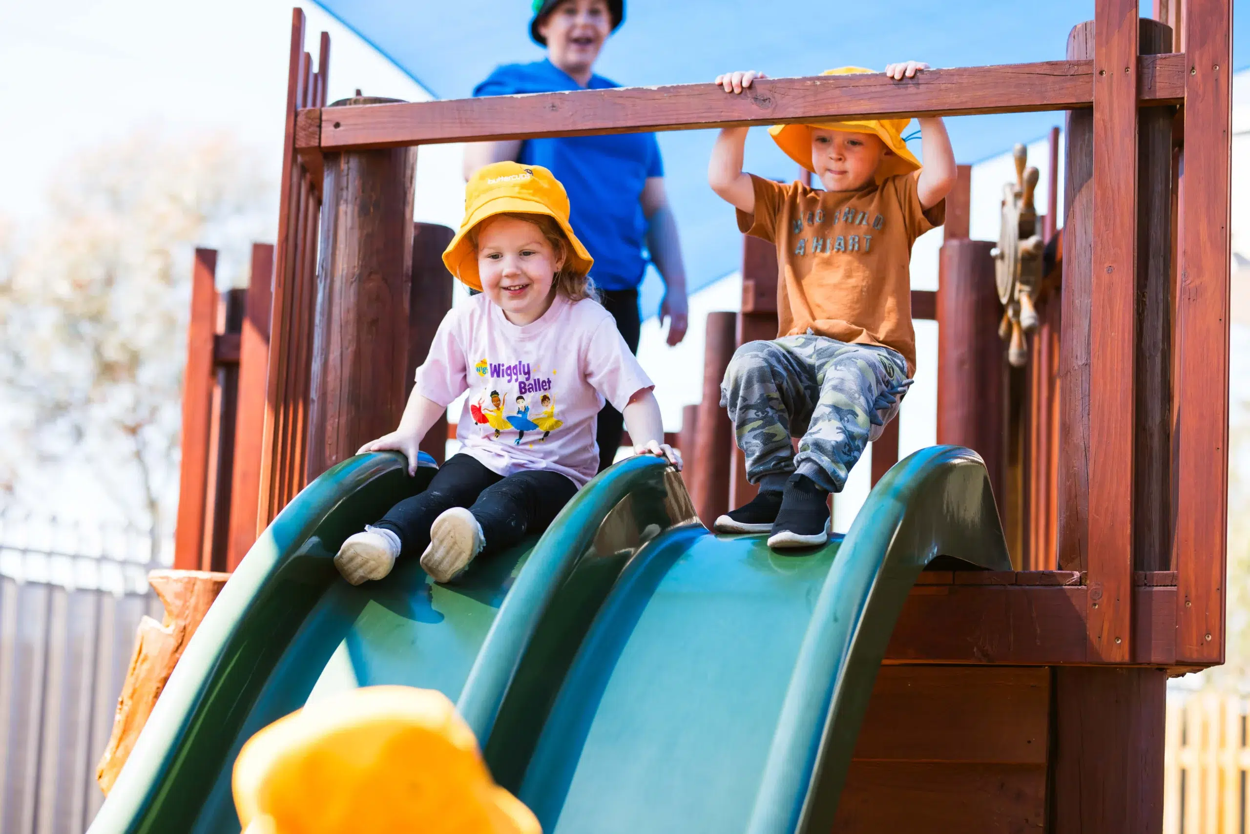 Children having fun sliding down green slides on a wooden playground at Buttercups Perth Childcare, with a staff member smiling in the background under a sunny blue sky.