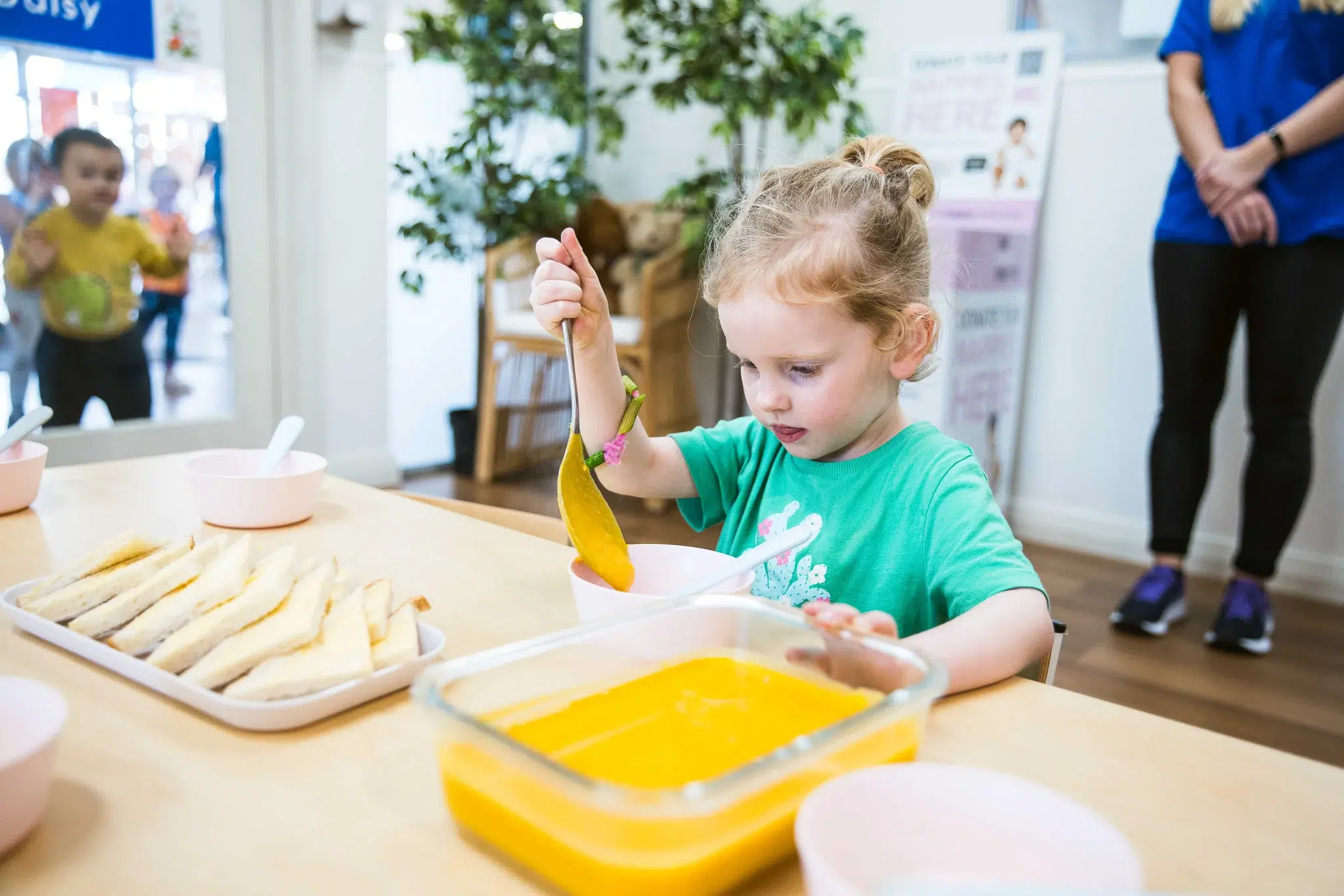 A young child at Buttercups Perth Childcare, wearing a green shirt, carefully ladles warm soup into a bowl during mealtime. The setting is a bright and welcoming dining area with a plate of neatly arranged sandwiches and child-friendly tableware, fostering independence and engagement in healthy eating.