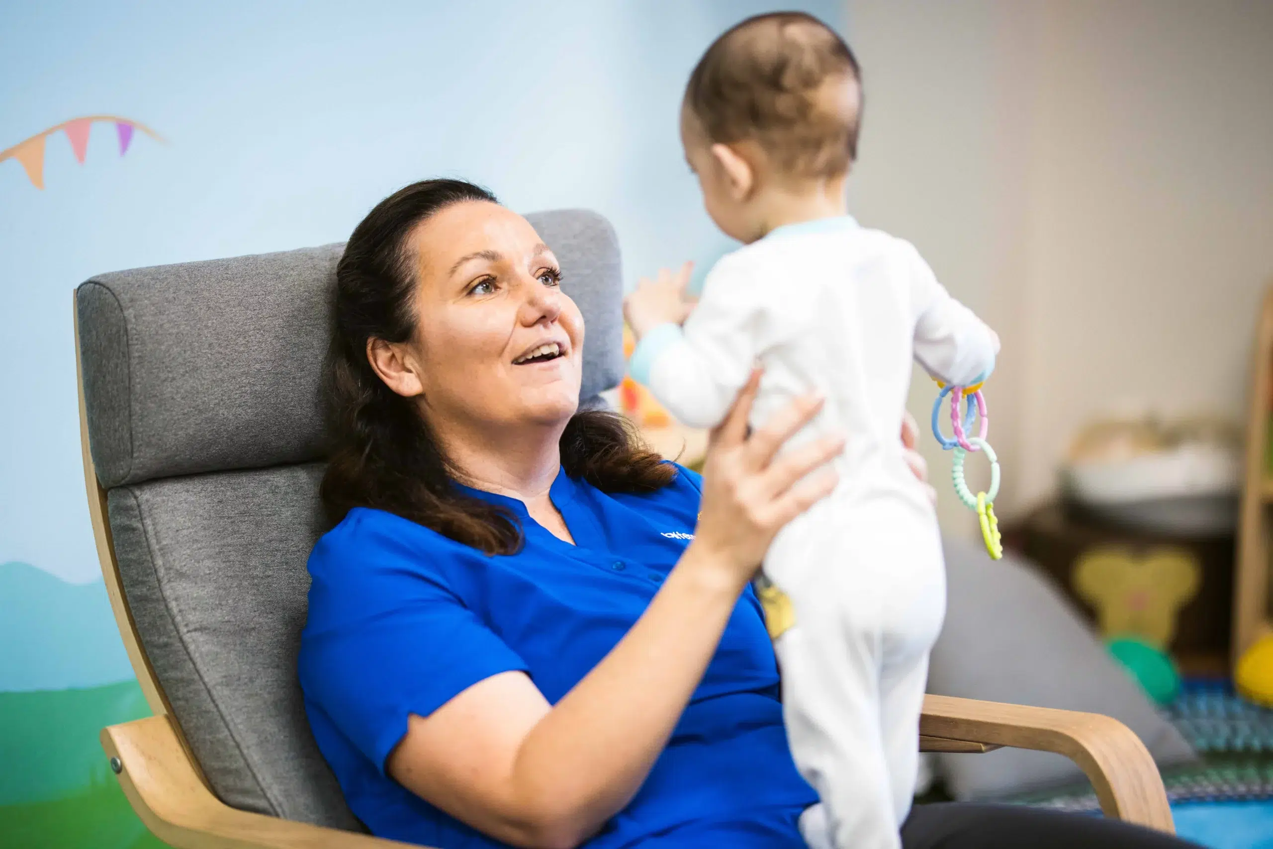 A warm and nurturing moment at Buttercups Perth Childcare, where an educator in a blue uniform sits in a cosy armchair, engaging with a baby dressed in white. The baby holds colourful teething rings while reaching out, highlighting the caring and interactive environment provided for early learning and development.