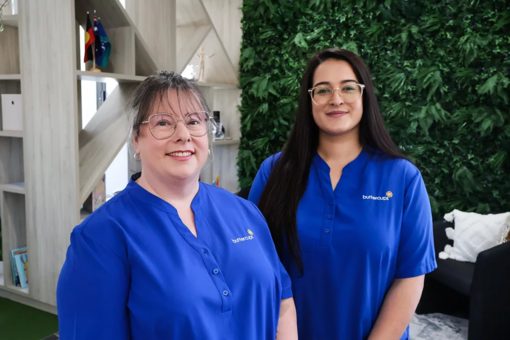 Buttercups Perth Childcare Koondoola staff members, wearing blue uniforms, standing in front of a greenery wall and wooden shelves, showcasing their welcoming and professional environment.