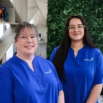 Buttercups Perth Childcare Koondoola staff members, wearing blue uniforms, standing in front of a greenery wall and wooden shelves, showcasing their welcoming and professional environment.