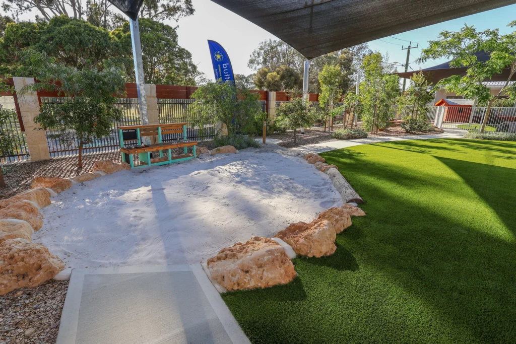 Buttercups Perth Childcare outdoor play area featuring a spacious sandpit surrounded by natural rocks, with a mud kitchen for imaginative play and sensory exploration.