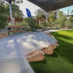 Buttercups Perth Childcare outdoor play area featuring a spacious sandpit surrounded by natural rocks, with a mud kitchen for imaginative play and sensory exploration.